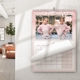 A pink glitter-bordered A3 calendar hanging on a white wall. The visible page shows a landscape photo of two people by a mountain lake. The lower portion reveals a partially turned monthly grid. A modern room with a leather sofa and a potted plant is in the background.