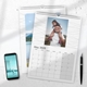 Two A4 photo calendars for 2025, opened to May and July. The July calendar features your own image. A smartphone and pen are placed next to the calendars on a white desk, with space for notes in the month grids.