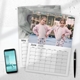 June 2026 wall calendar lying on a surface, featuring a photo of twin girls in matching outfits at a fairground. A second calendar underneath shows part of May 2026 with a black-and-white close-up image. Also visible are a smartphone and a black pen placed nearby.