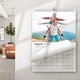 August 2026 wall calendar hanging on a white wall in a modern room. The calendar features a joyful photo of a father giving a piggyback ride to two children in a mountain landscape, all posing with arms outstretched like aeroplanes. The calendar page is partially turned, revealing clean date boxes below.