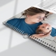 Close-up view of a wall calendar lying flat, showcasing a tender moment between a mother in a blue shirt and her baby lying beside her. The image highlights the sturdy wire binding and metal hanging hook, with part of the calendar grid visible in the top corner.