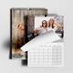 Two wall calendars displayed on a light surface. The front calendar shows a cheerful photo of two women in sunglasses outdoors, with the page partly turned to reveal the monthly grid below. The second calendar in the background features a cosy indoor scene with a smiling woman and festive fairy lights. Both calendars are spiral-bound with a hanging hook.