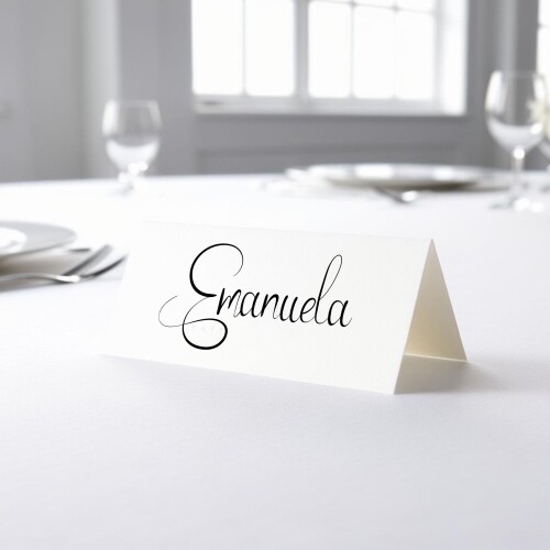 Personalised wedding place card with your name in elegant black script on folded white cardstock. Placed on a table set for a formal event, with plates, cutlery, and wine glasses in the softly blurred background.