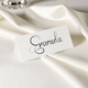 Personalised wedding place card with your name in elegant black script on folded white cardstock. Positioned on a neatly folded white napkin atop a white plate, with a soft-focus white rose in the background.