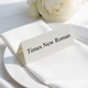 Personalised wedding place card with your name in elegant black script on folded white cardstock. Positioned on a neatly folded white napkin atop a white plate, with a soft-focus white rose in the background.