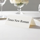 Personalised wedding place card with your name in elegant black script on folded white cardstock. Displayed on a white tablecloth with wine glasses, plates, and a floral centrepiece in the softly blurred background.