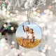 Personalised ceramic bauble featuring two children sitting on a haystack. The older child points into the distance while smiling, with a snowy Christmas tree branch and festive bokeh lights in the background.