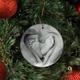 Personalised ceramic bauble hanging on a Christmas tree, featuring a black-and-white image of adult hands forming a heart shape around a baby's feet. Red baubles and pine branches add a festive touch.