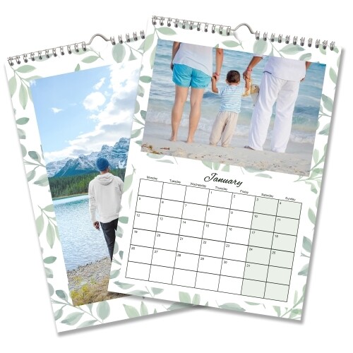 A 2026 wall calendar with a soft green leaf border design is shown open to January. The top image features a family standing barefoot at the beach, holding hands. The calendar grid below displays the month of January with clearly marked dates. Behind it, another page shows a man in a white hoodie and blue hat standing by a mountain lake with snowy peaks in the distance. The calendar is spiral-bound with a central hanging loop.