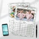 Two 2026 wall calendars with a green leaf border are displayed on a white surface. The top calendar is open to April, featuring a photo of twin girls in matching pink jackets posing confidently in front of a carousel. The calendar grid shows all days of the month. The calendar underneath displays a colourful toy photo. A smartphone and black pen rest nearby, creating a clean, organised workspace scene.