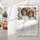 A 2026 wall calendar hangs on a light-coloured wall in a bright, modern room. It is open to April, featuring a photo of two children in white shirts—one wearing a straw hat—posing in front of pastel balloons. The page is being turned, partially revealing the month’s grid. The calendar has a soft green leaf border and is spiral-bound with a central hanging hook. A stylish lounge area is visible in the background.