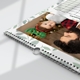 A close-up of a 2026 wall calendar lying flat on a white surface, featuring a photo of a man holding a baby wearing festive green reindeer antlers in front of a Christmas tree. The calendar has a soft green leaf border and a visible spiral binding with a central metal hanging loop, highlighting its durable and practical design. The corner of the December calendar grid is partially visible.
