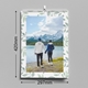 A 2026 wall calendar measuring 210mm wide by 297mm tall is displayed against a grey wall. The cover features a couple holding hands, standing beside a lake with snow-capped mountains and forest in the background. The calendar has a soft green leaf border, spiral binding with a central hanging hook, and is shown in a vertical A4 portrait format.