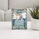 A Christmas card featuring a family of four dressed casually, with two children standing between their smiling parents. The card is decorated with a festive border of holly, pinecones, and greenery. The message reads, 