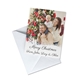 A personalised Christmas card featuring a smiling family of three, with the man holding a baby on his shoulders and the woman standing beside them. The background shows a decorated Christmas tree, and the card is placed on a white envelope. It reads, 