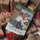 A personalised Christmas card featuring a smiling man with two children outdoors. The boy is on his shoulders while the girl stands beside them. The card, surrounded by wrapped presents and festive decorations, reads, 