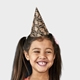 A young girl wearing a personalised party hat featuring a repeated print of her own face with a golden headband. The girl is smiling widely, and the hat perfectly matches her cheerful expression. The background is plain white.