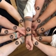 A group of people standing in a circle, each placing their forearm inward to form a star shape with hands linked. Each person has a black circular temporary tattoo on their forearm that reads Sally's Hen Party with a small face graphic.