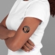 A woman with crossed arms wearing a white T-shirt displays a black circular temporary tattoo on her upper arm. The tattoo reads Sally's Hen Party Brighton 2025 and includes a smiling woman's face in the centre.