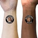 Two wrists, one light-skinned and one dark-skinned, each displaying a black circular personalised temporary tattoo with a woman's face and the text Sally's Hen Party Brighton 2025. The tattoos offer a personalised touch for a hen party celebration.