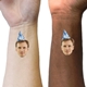 Two wrists, one light-skinned and one dark-skinned, both display temporary tattoos featuring a man's face wearing a blue party hat marked with 