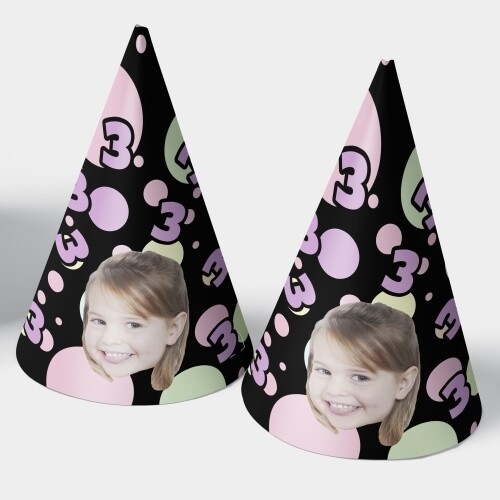 Pair of black personalised party hats featuring a smiling young girl's face, decorated with pastel pink, green, and purple bubbles and the number 3, ideal for a third birthday celebration.