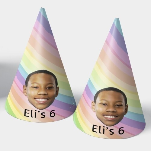 Two customised party hats featuring a rainbow stripe design, a personalised photo of a smiling boy, and the text Eli's 6. The hats are displayed upright, perfect for celebrating a 6th birthday or rainbow-themed parties.