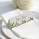 Personalised wedding place card with your name in elegant design on folded white cardstock. Positioned on a neatly folded white napkin atop a white plate, with a soft-focus white rose in the background.