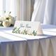 Personalised wedding place card with your name in elegant design on folded white cardstock. Displayed on a table with a flowing white tablecloth, with soft natural light and white roses in the background.