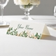 Personalised wedding place card with your name in elegant design on folded white cardstock. Displayed on a white tablecloth with wine glasses, plates, and a floral centrepiece in the softly blurred background.