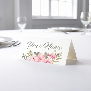 Personalised wedding place card with your name in elegant design on folded white cardstock. Placed on a table set for a formal event, with plates, cutlery, and wine glasses in the softly blurred background.