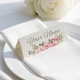 Personalised wedding place card with your name in elegant design on folded white cardstock. Positioned on a neatly folded white napkin atop a white plate, with a soft-focus white rose in the background.