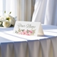 Personalised wedding place card with your name in elegant design on folded white cardstock. Displayed on a table with a flowing white tablecloth, with soft natural light and white roses in the background.