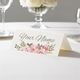 Personalised wedding place card with your name in elegant design on folded white cardstock. Displayed on a white tablecloth with wine glasses, plates, and a floral centrepiece in the softly blurred background.