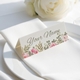 Personalised wedding place card with your name in elegant design on folded white cardstock. Positioned on a neatly folded white napkin atop a white plate, with a soft-focus white rose in the background.