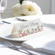 Personalised wedding place card with your name in elegant designt on folded white cardstock. Placed on a white plate with silver cutlery and a neatly folded napkin, with soft-focus white flowers in the background.