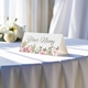 Personalised wedding place card with your name in elegant design on folded white cardstock. Displayed on a table with a flowing white tablecloth, with soft natural light and white roses in the background.