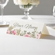 Personalised wedding place card with your name in elegant design on folded white cardstock. Displayed on a white tablecloth with wine glasses, plates, and a floral centrepiece in the softly blurred background.