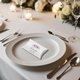 Personalised wedding favour pillow box with a floral pink rose design placed on a white dinner plate. The elegant table setting includes silver cutlery, wine glasses, and candlelight, with white roses in the background, creating a romantic atmosphere.