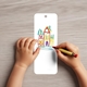 Child drawing a colourful house with crayons on a blank white bookmark with rounded corners and a top hole. The bookmark rests on a light wooden surface, ideal for personalised kids crafts, school projects or custom creative activities.