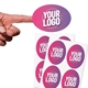 A hand holding a personalised oval sticker with a pink-to-purple gradient and placeholder text Your Logo. Below, two sheets of matching custom stickers are displayed, each featuring multiple identical labels. Ideal for branding, product packaging, or personalised use.