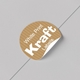 Round personalised Kraft label with white print partially peeled from a smooth grey surface. The custom sticker features a brown striped texture and bold white text reading White Print Kraft Labels, highlighting its easy-peel design and suitability for packaging or branding.