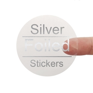 A transparent square sticker with silver foil text reading Silver Foiled Stickers. A person's finger is visible behind the sticker, showing its clear background. The silver foil lettering has a reflective metallic finish for a premium look.