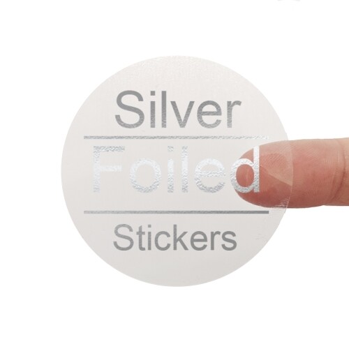 A transparent square sticker with silver foil text reading Silver Foiled Stickers. A person's finger is visible behind the sticker, showing its clear background. The silver foil lettering has a reflective metallic finish for a premium look.