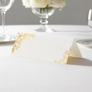 wedding place card with an elegant design on folded white cardstock ready to write your name on. Displayed on a white tablecloth with wine glasses, plates, and a floral centrepiece in the softly blurred background.