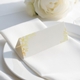 wedding place card with an elegant design on folded white cardstock, ready to write your names on. Positioned on a neatly folded white napkin atop a white plate, with a soft-focus white rose in the background.