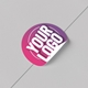 Close-up of a round sticker with the text Your Logo in white on a pink and purple background, partially peeling from a smooth grey surface.