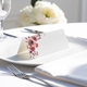 blank wedding place card with elegant design on folded white cardstock. Placed on a white plate with silver cutlery and a neatly folded napkin, with soft-focus white flowers in the background.