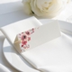 wedding place card with an elegant design on folded white cardstock, ready to write your names on. Positioned on a neatly folded white napkin atop a white plate, with a soft-focus white rose in the background.