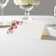 wedding place card with an elegant design on folded white cardstock ready to write your name on. Displayed on a white tablecloth with wine glasses, plates, and a floral centrepiece in the softly blurred background.